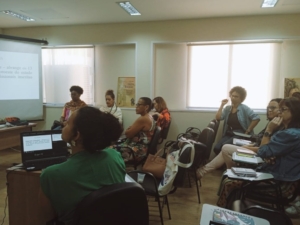 #DescriçãoDaImagem Na imagem, aparecem conselheiras do CRESS-RJ assistindo à apresentação da Seccional Norte Fluminense. A conselheira Ully Azevedo, da Seccional, uma mulher parda vestindo blusa verde, está sentada com um laptop em uma bancada a sua frente fazendo a apresentação. As outras conselheiras olham par Ully e assistem à apresentação. Há cadeiras azuis espalhadas no auditório e as paredes são claras.